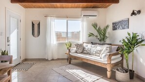 Een gezellige woonkamer met een houten bank, een groot raam met gordijnen en verschillende potplanten.
