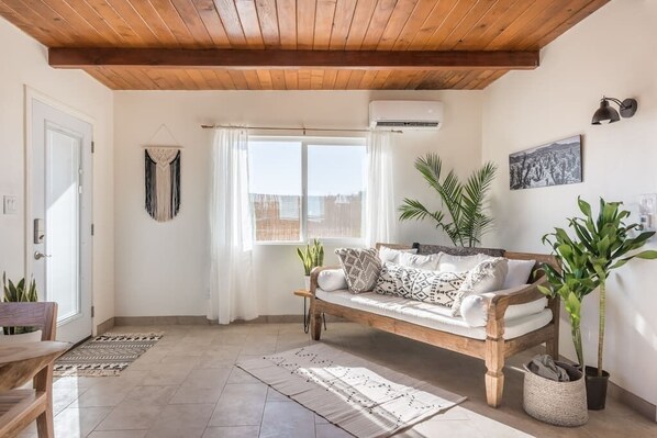 A cozy living room with a wooden bench, a large window with curtains, and several potted plants.