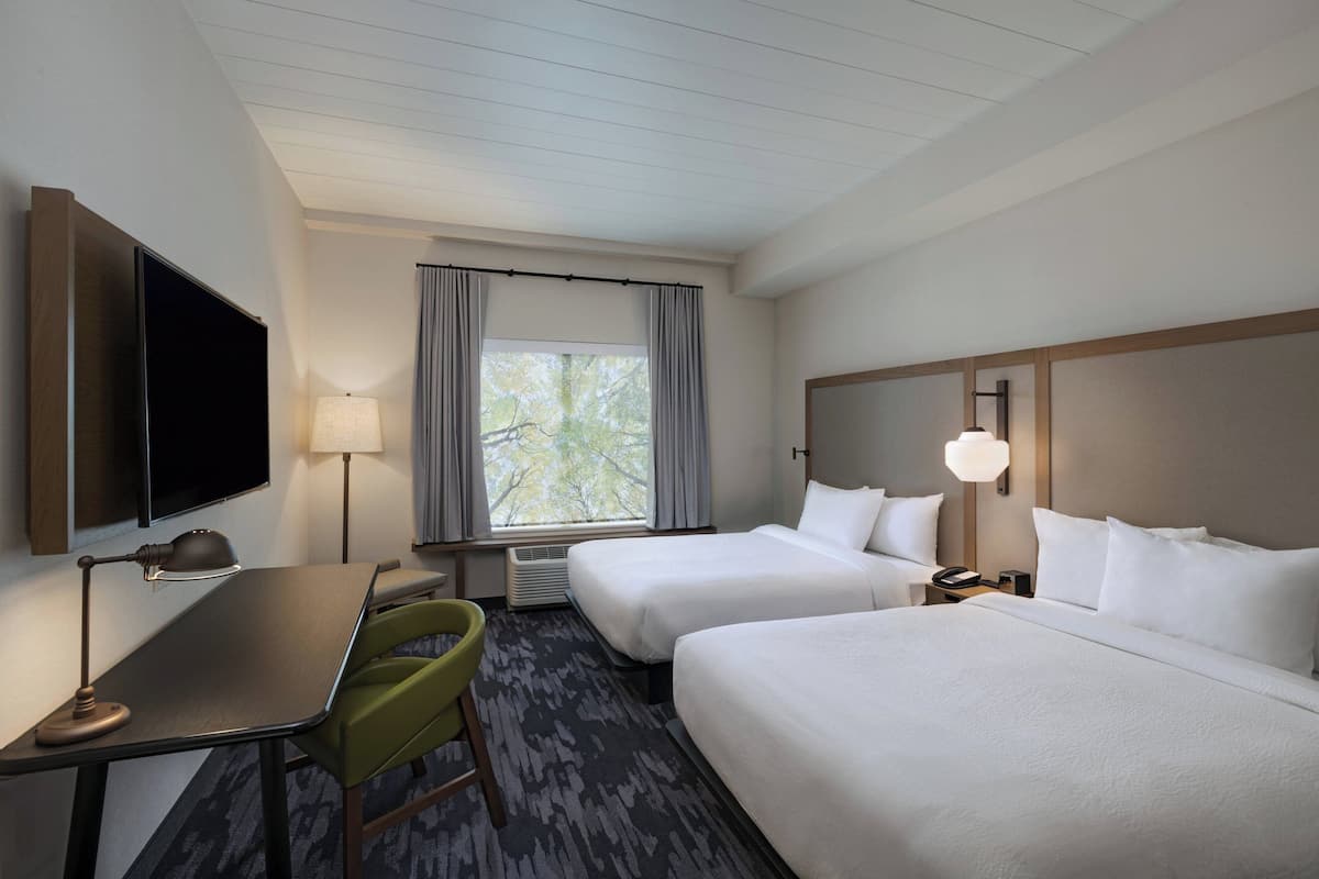 A hotel room with two beds, a desk, a chair, a TV, a lamp, and a window with curtains.