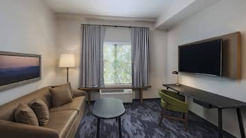 A hotel room with a sofa, a small table, a desk, a TV, a window with curtains, and an air conditioner.