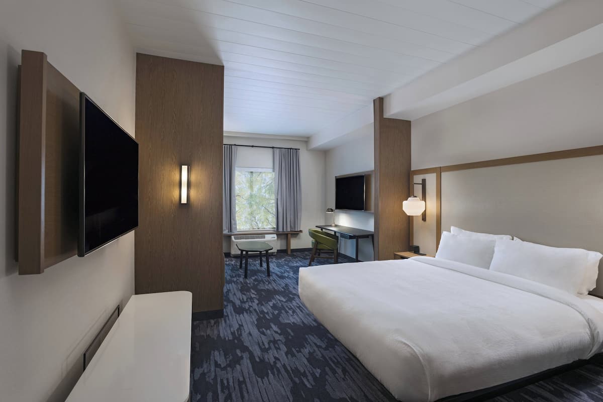 A hotel room with a large bed, a desk, a chair, a flat-screen TV mounted on the wall, and a window with curtains.