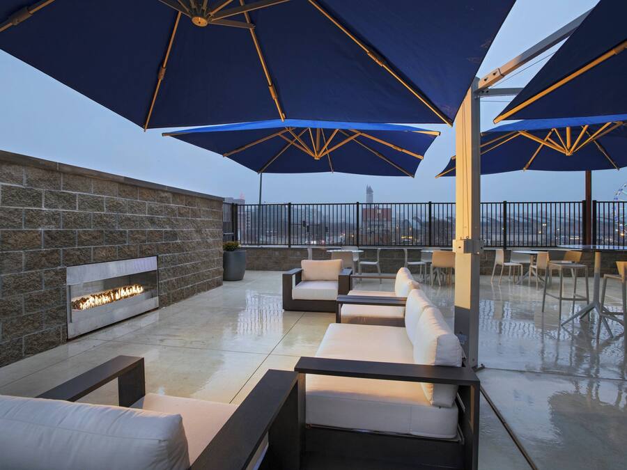 An outdoor seating area with multiple tables and chairs, large umbrellas, and a built-in fire feature against a stone wall.
