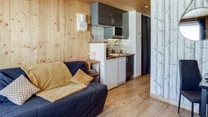 A cozy room with a wooden ceiling, a dark blue sofa with a yellow throw, a small table with a chair, and a kitchen area with a microwave and cabinets. - Le Curling A (Val Claret) by Interhome (Tignes)