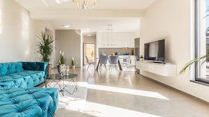 A modern living room with a teal sofa, a glass coffee table, and a TV. There is a kitchen in the background with white cabinets and a dining area. - Luxurious home in Glavina Donja (Glavina Donja)
