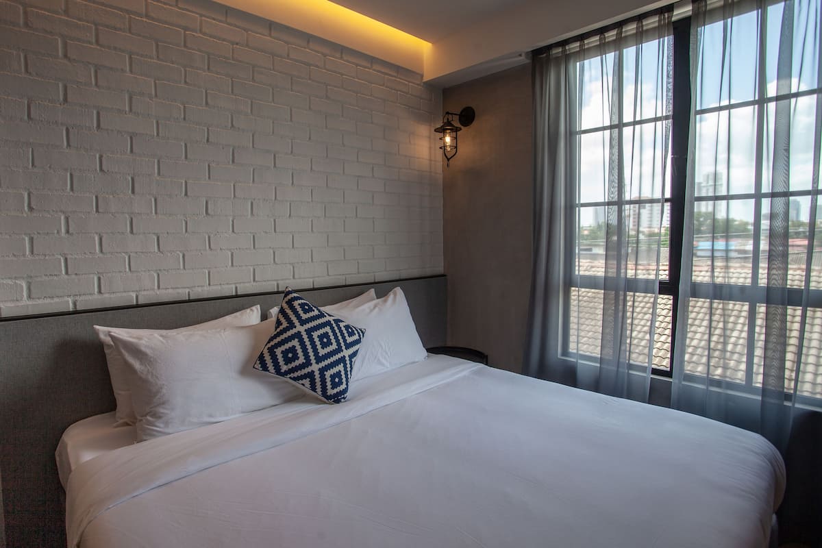 A hotel room with a white brick wall, a bed with white bedding and a patterned pillow, a window with sheer curtains, and a wall-mounted light fixture.