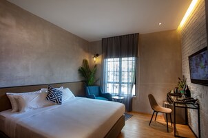 Una habitación de hotel moderna con una cama grande, un escritorio con silla, una mesita pequeña y un amplio ventanal con cortinas.