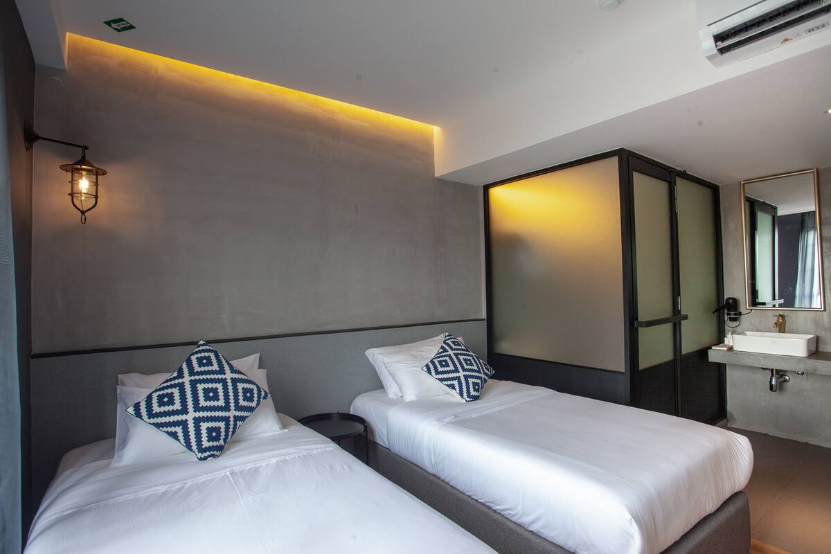 A hotel room with two beds, a bedside table, a mirror, and a bathroom with a sink and mirror.