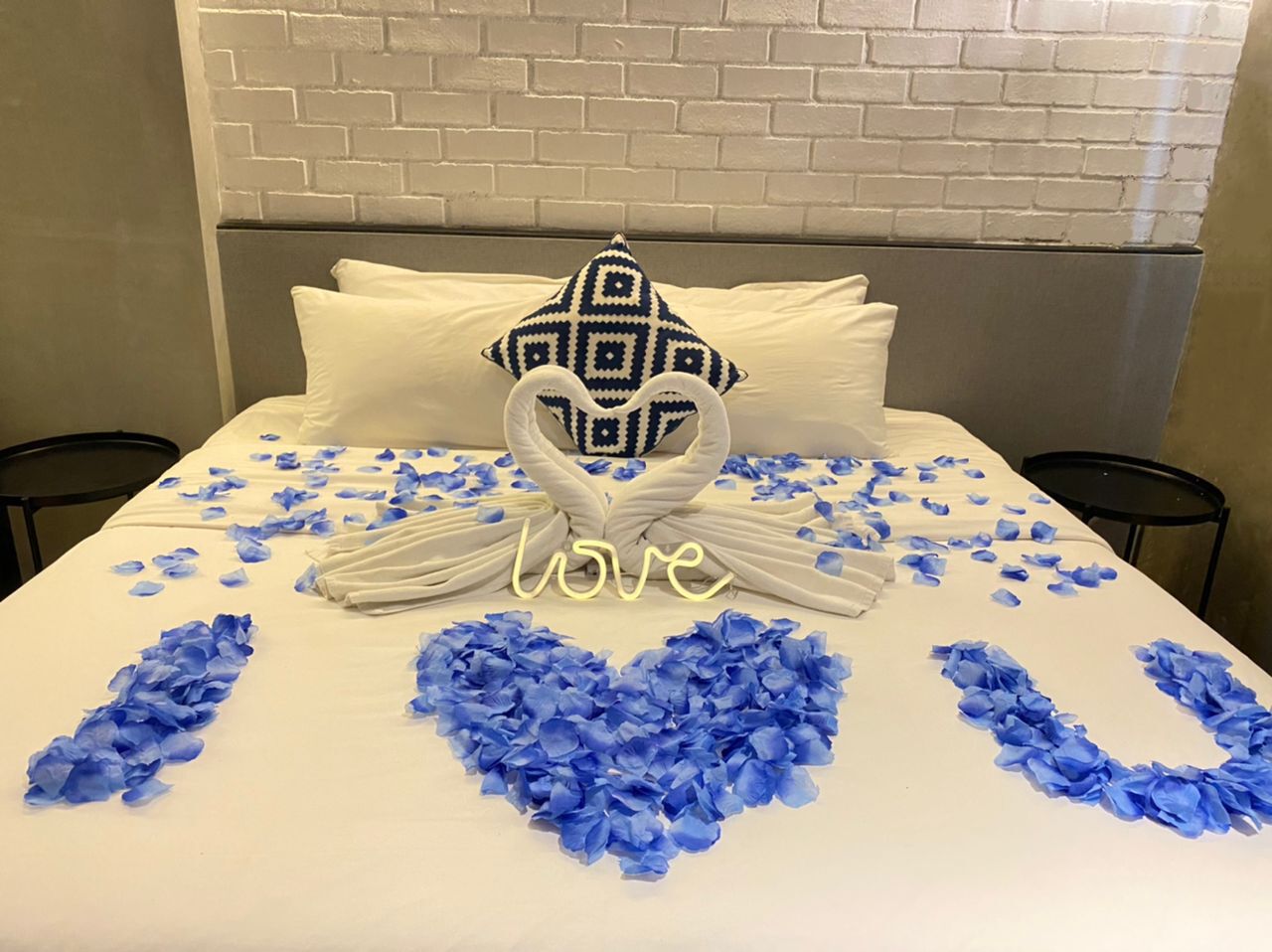 Una cama con cubrecama blanco, un cojín decorativo y pétalos de flores azules dispuestos en forma de corazón con la palabra "love" escrita.