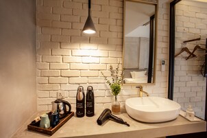Une salle de bain avec un mur en briques blanches, un grand miroir, un lavabo blanc, un sèche-cheveux et deux bouteilles d’eau réutilisables.