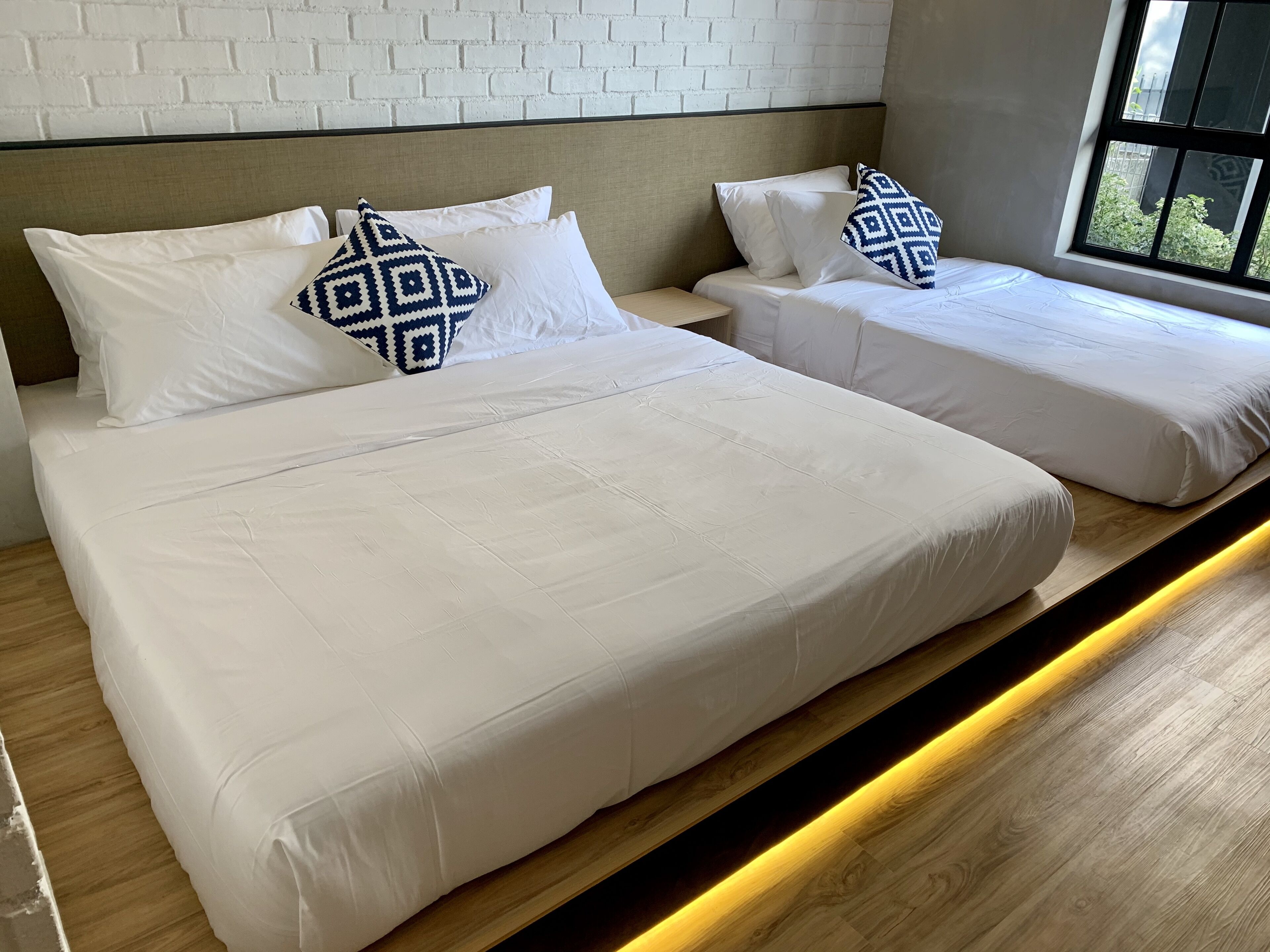 A hotel room with two beds, white bedding, patterned pillows, a wooden floor, and a window with a view of greenery.