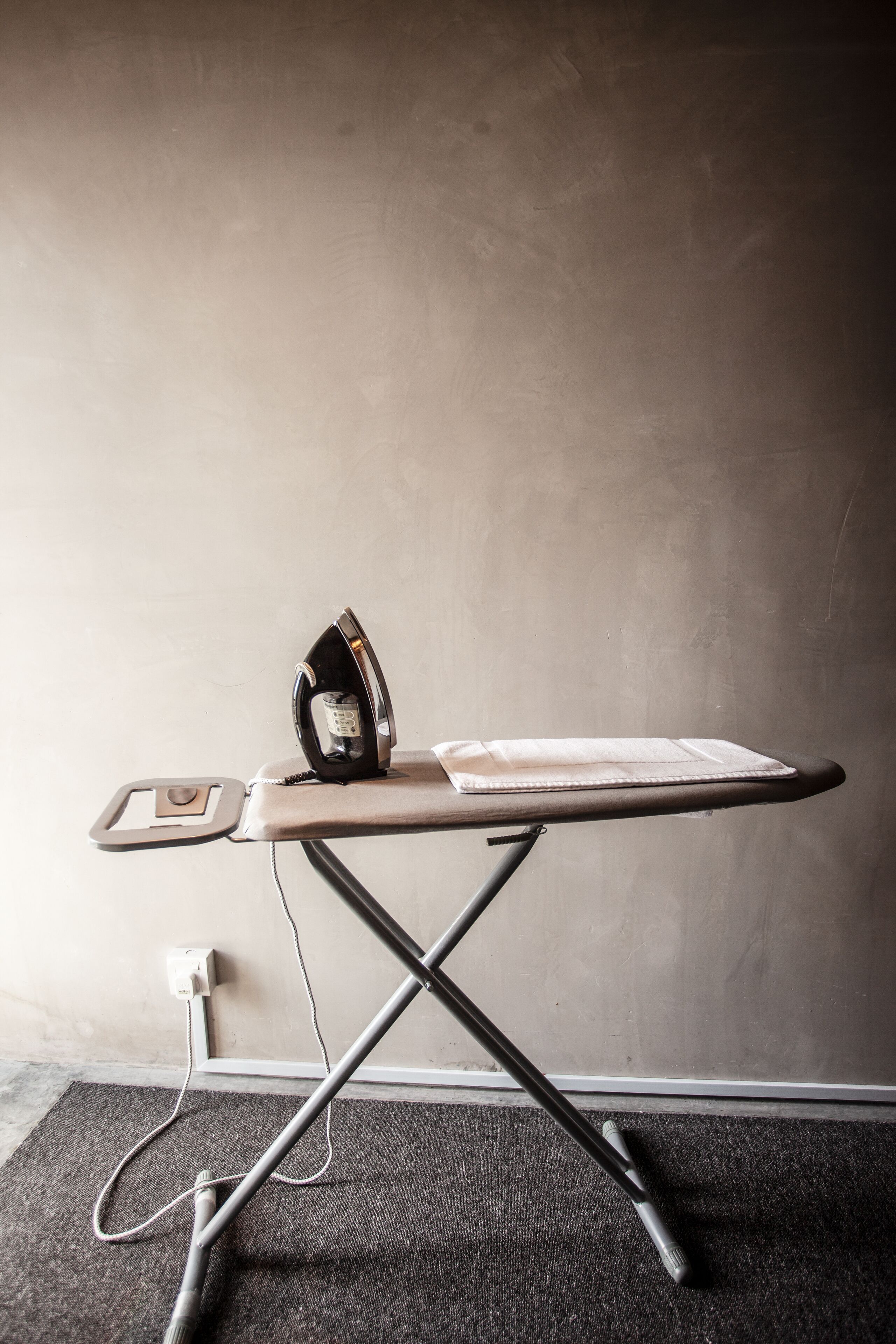 Une planche à repasser avec un fer à repasser et un morceau de tissu plié, une prise de courant et un sol recouvert de moquette.