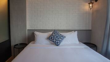 Una cama bien hecha con sábanas blancas y un cojín estampado en azul, una pared de ladrillo blanco y un aplique en la pared.