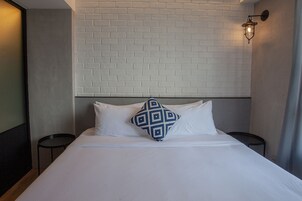 A neatly made bed with white linens and a blue patterned pillow, a white brick wall, and a wall-mounted light fixture.