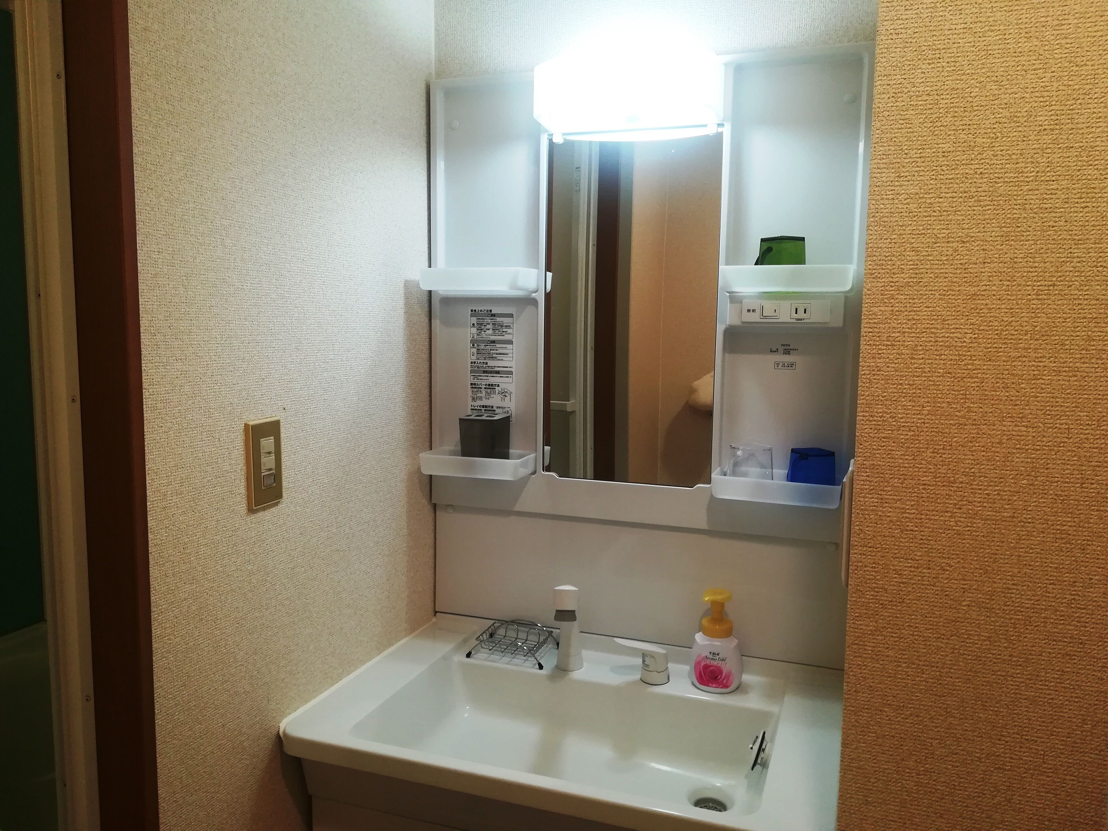 Un baño con lavamanos blanco, un espejo, dispensador de jabón y una pequeña estantería con artículos de tocador.