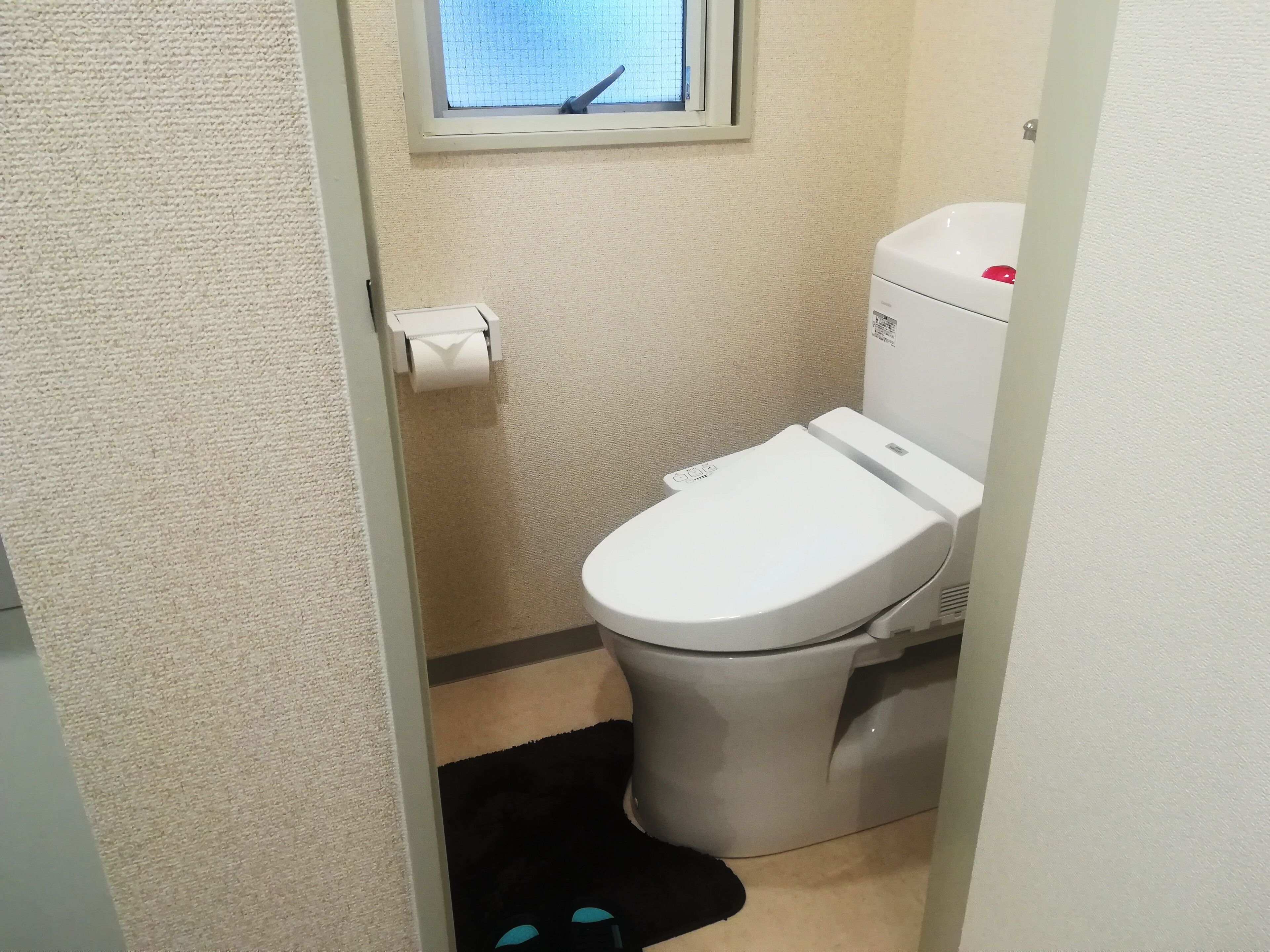 A small bathroom with a toilet, a window, and a roll of toilet paper.