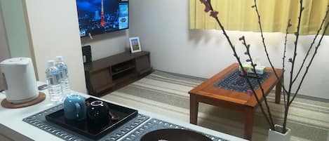Una sala moderna con televisor de pantalla plana, cafetera, botellas de agua, una bandeja con bolsitas de té y una mesita con un florero de ramas.