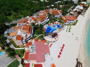 Vue aérienne d’un complexe hôtelier comprenant plusieurs bâtiments, une piscine centrale et une zone de plage avec des transats.