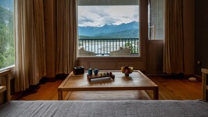 A room with a wooden floor, a bed, a table with a fruit bowl, and a view of mountains and a river through a window. - Songtsam Linka Bomi (Nyingtri)