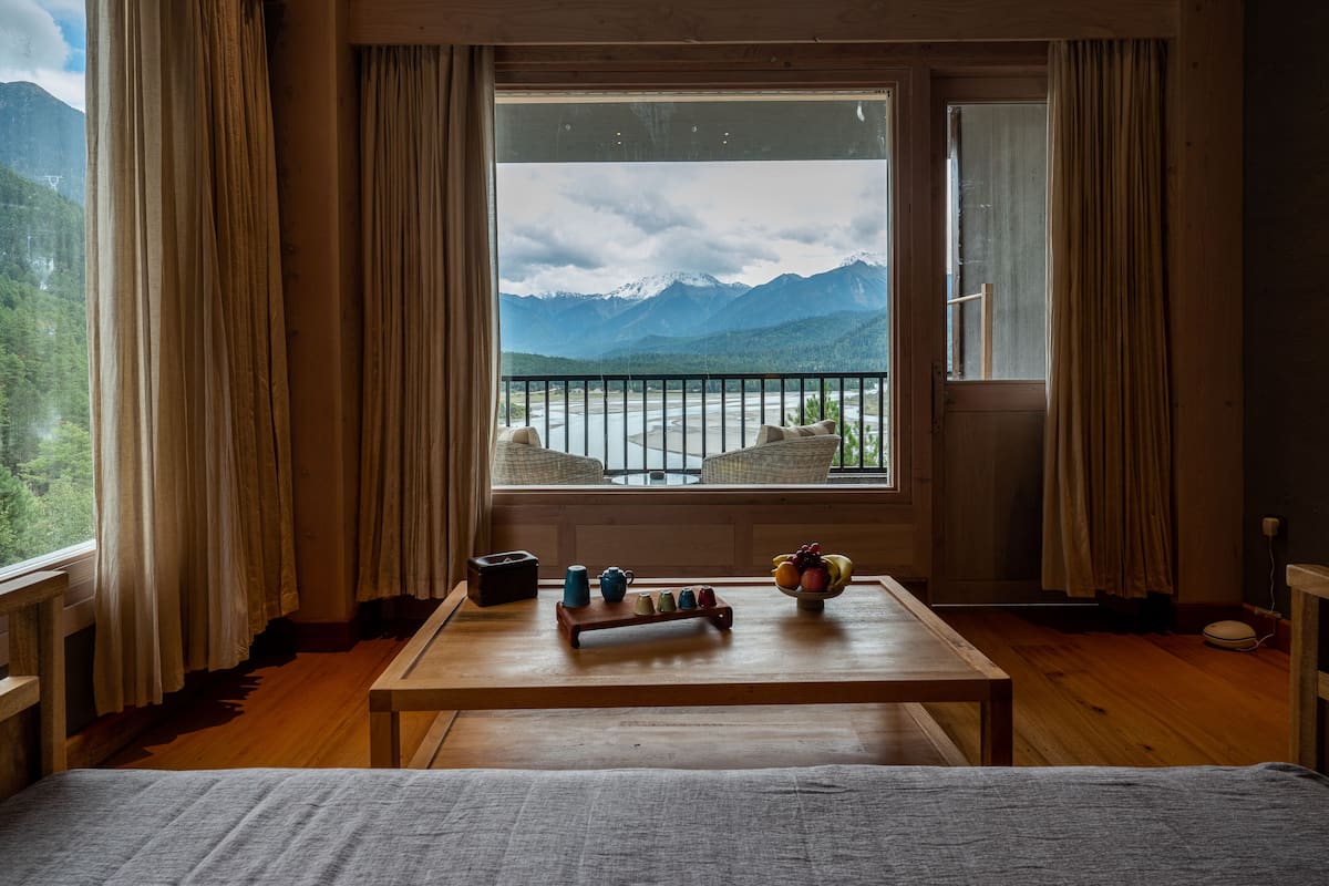 Quarto com piso de madeira, cama, mesa com tigela de frutas, vista para montanhas e rio pela janela.