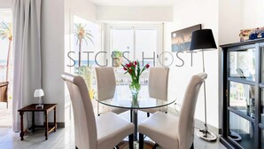A modern room with a glass table, four chairs, a small side table, a floor lamp, and a large window with a view of palm trees and the sea. - Beautiful Seafront Views (Sitges)