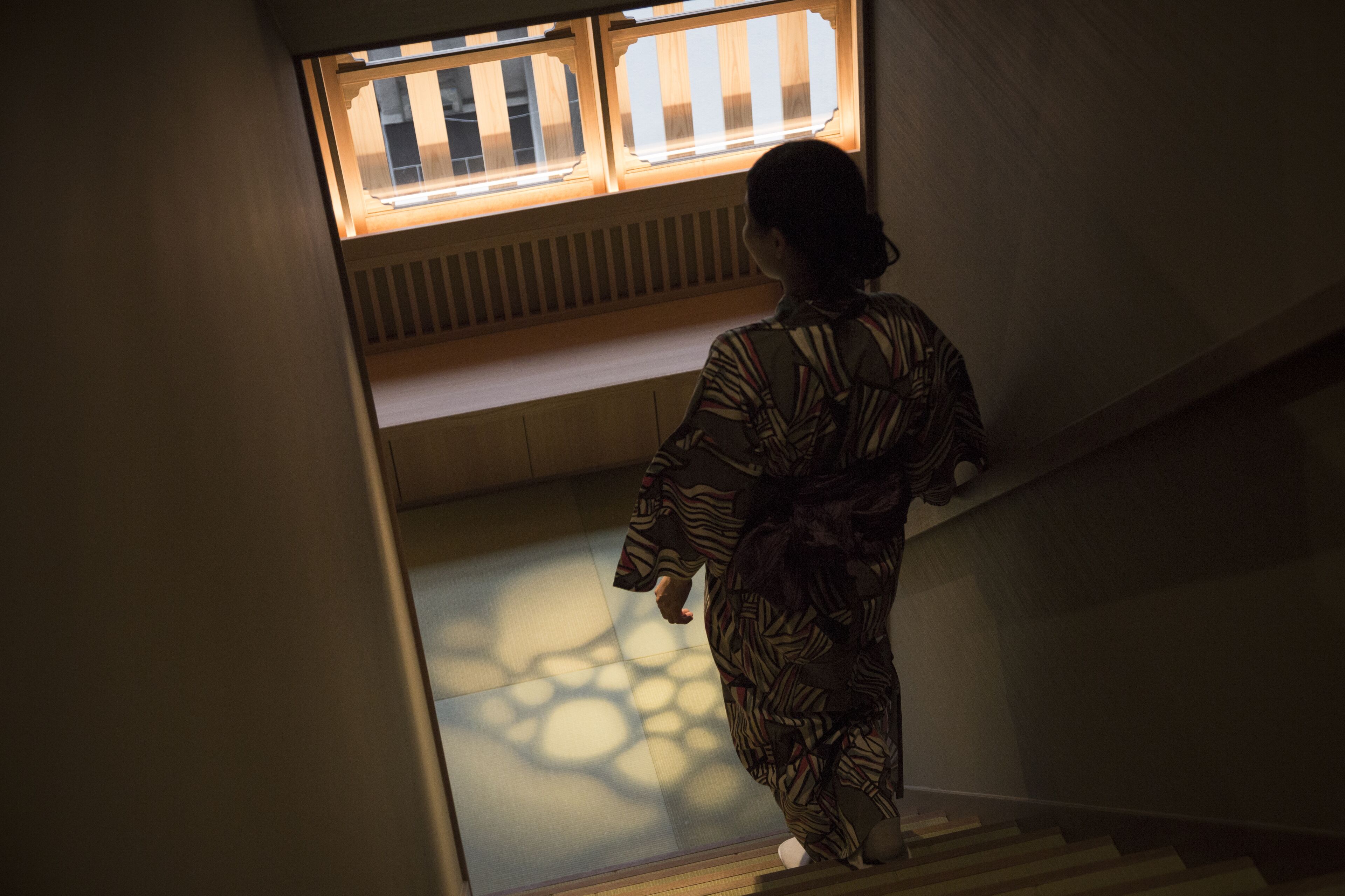A person descending a staircase in a building with a patterned floor and a window with vertical blinds.