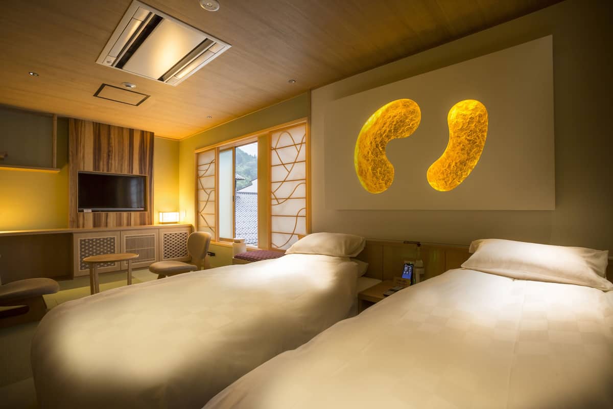 A hotel room with two beds, a TV, a desk, and a large illuminated artwork on the wall.
