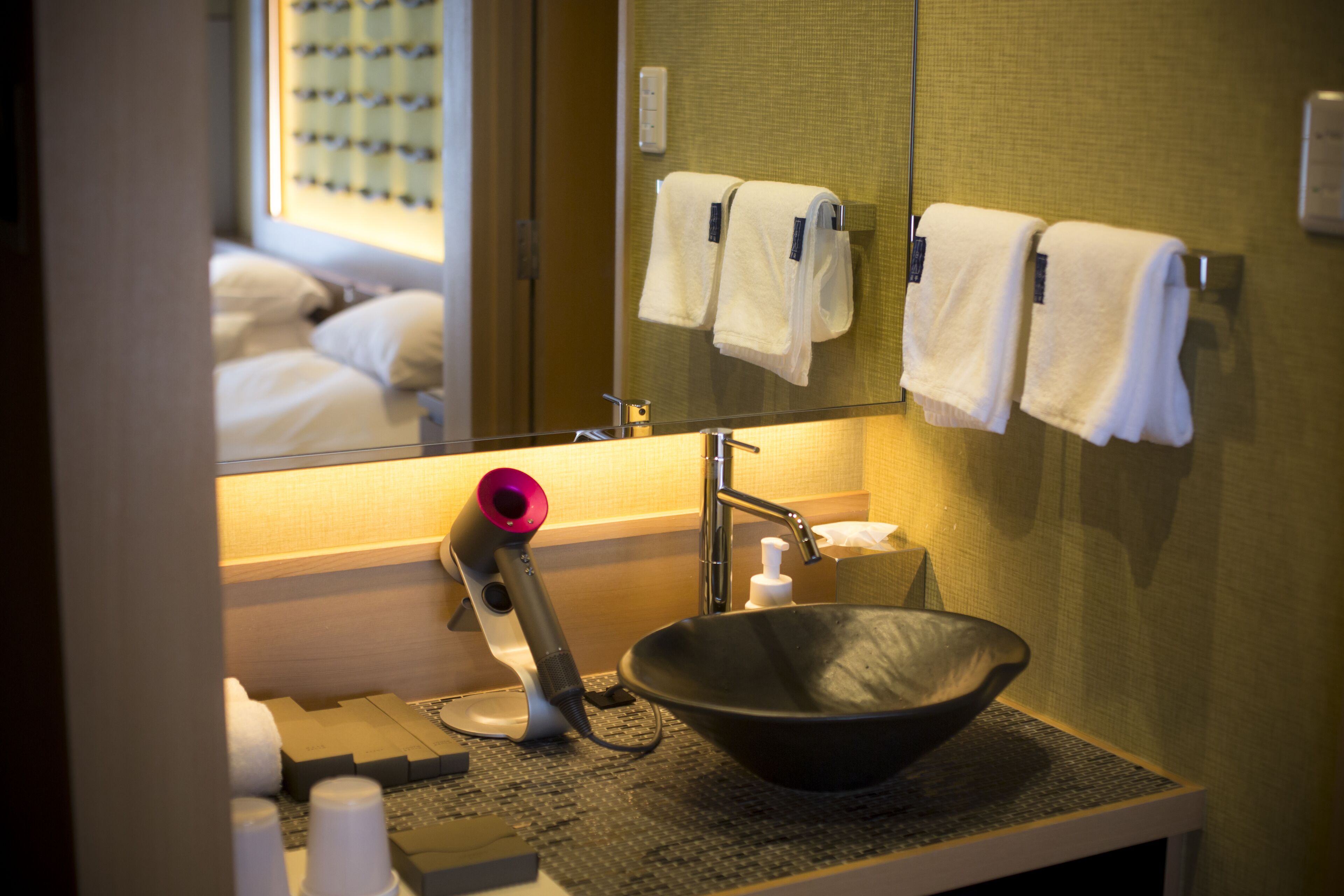 A hotel bathroom with a large mirror, a sink with a black bowl, a hairdryer, and towels hanging on a rack.