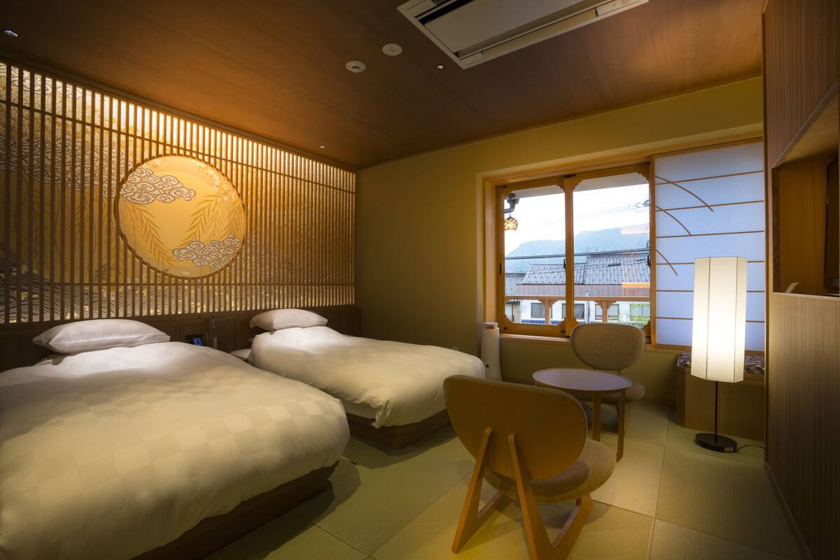 A hotel room with two beds, a small table, a chair, a lamp, and a view of buildings through a window.