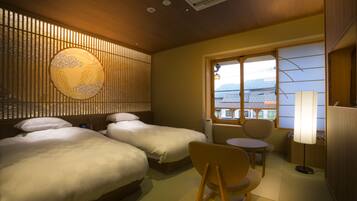 A hotel room with two beds, a small table, a chair, a lamp, and a view of buildings through a window.