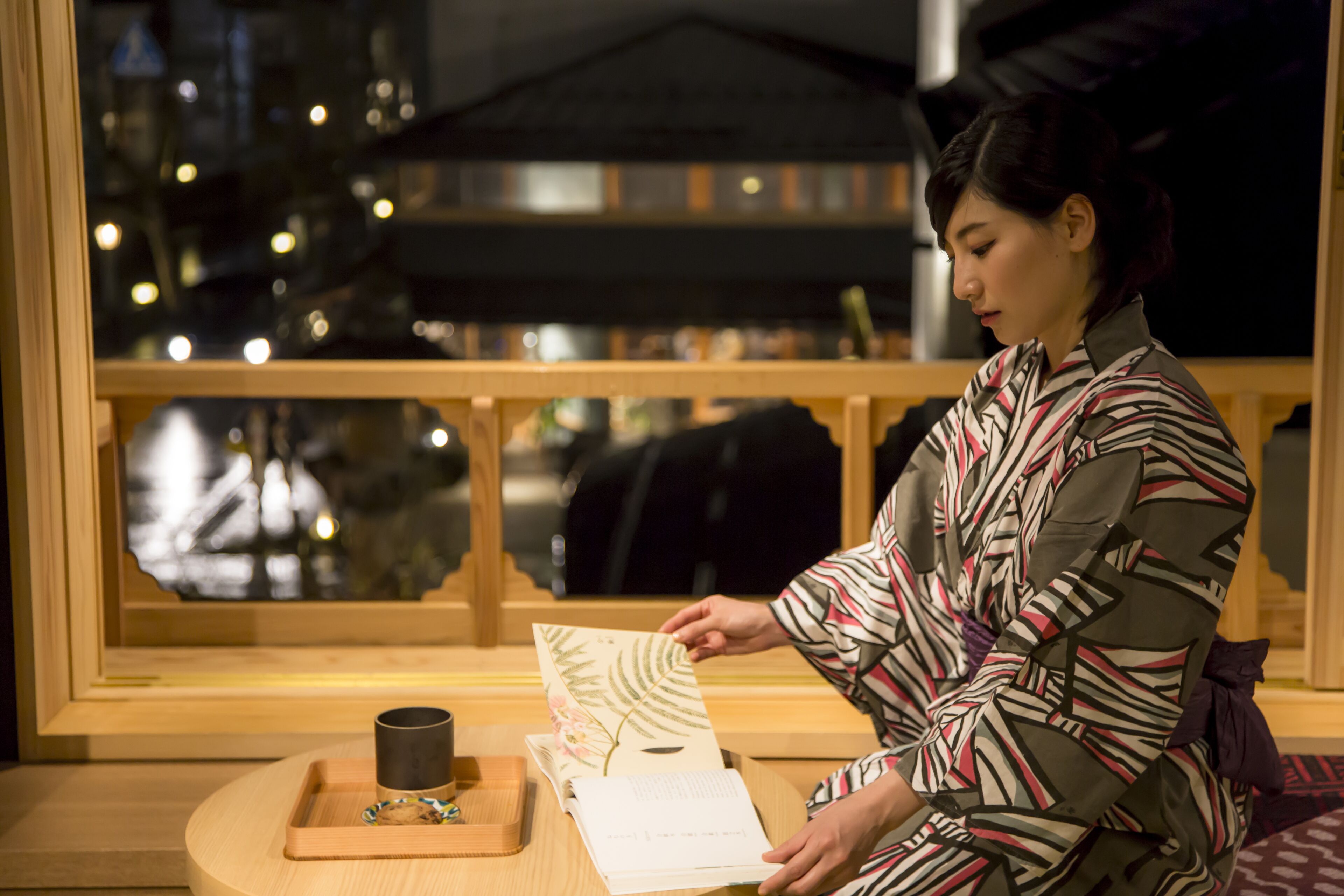 Una persona sentada junto a una ventana con vistas a un paisaje urbano, sosteniendo un libro y una taza sobre una bandeja de madera.