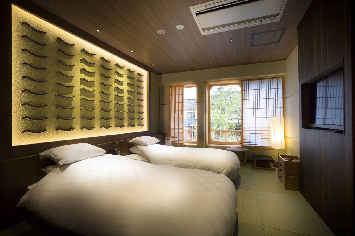 A hotel room with two beds, a wall-mounted shelf, a window with a view, and a small table.