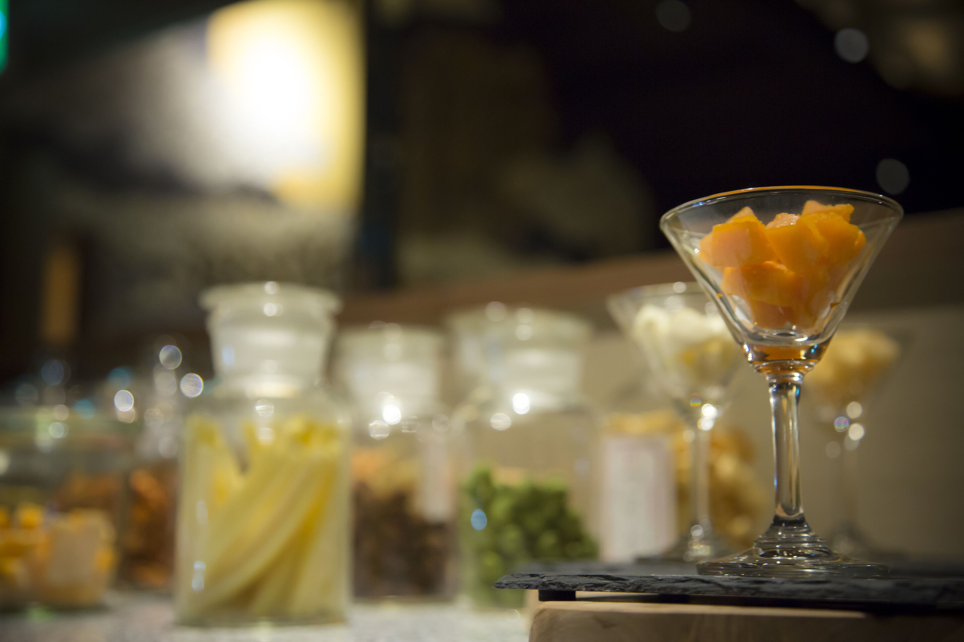 A glass with fruit on a bar counter, surrounded by jars of condiments.