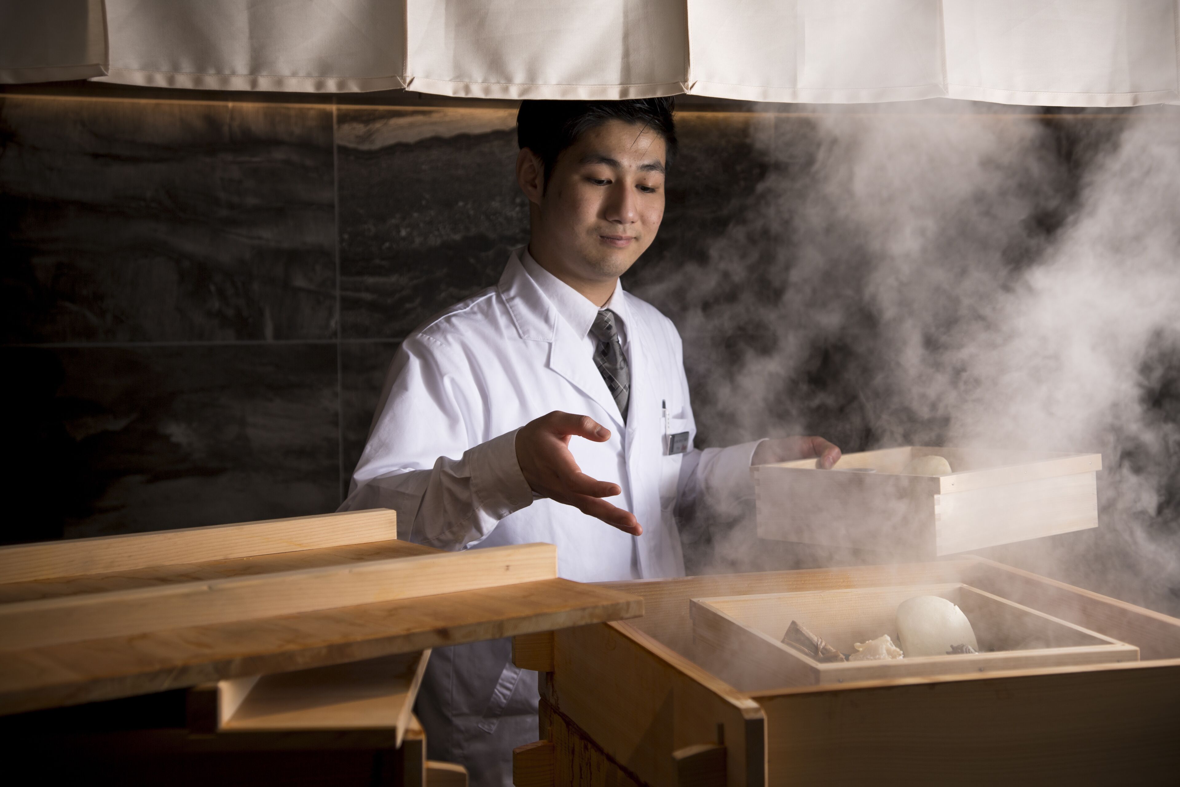 Um chef de cozinha com avental branco e gravata, em pé numa cozinha com bancada de madeira e uma caixa a emitir vapor.