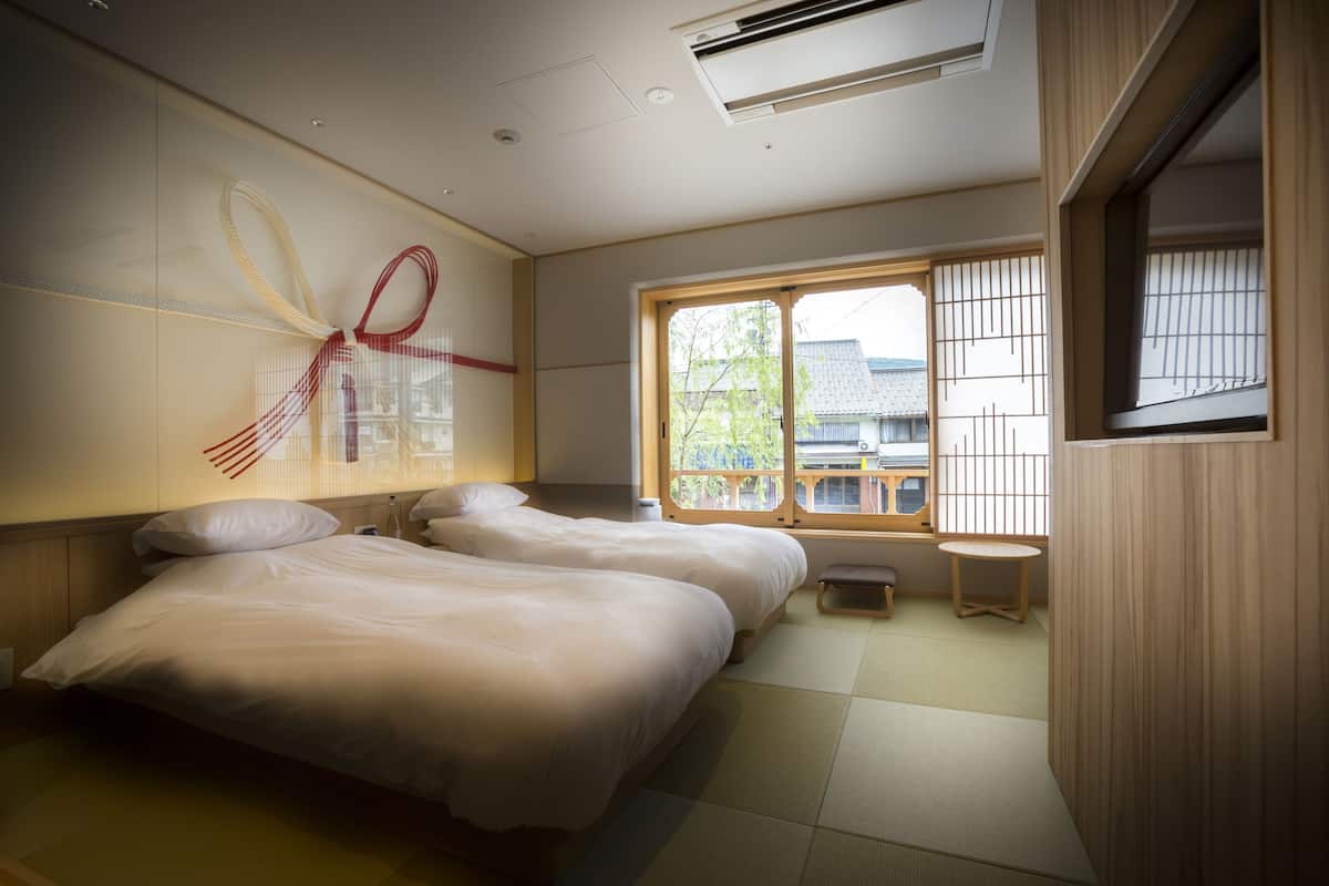 A hotel room with two beds, a small table, a window with a view of buildings, and a decorative wall piece.