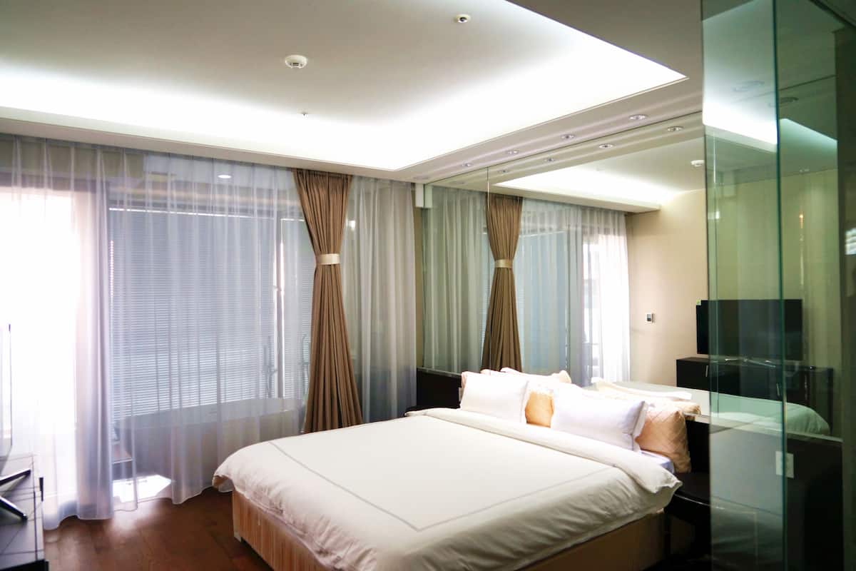 A modern hotel room with a large bed, a glass partition, and a view of the city through sheer curtains.