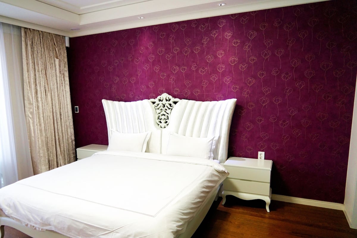 A bedroom with a large bed, a white headboard, a nightstand, and a purple floral wallpaper.