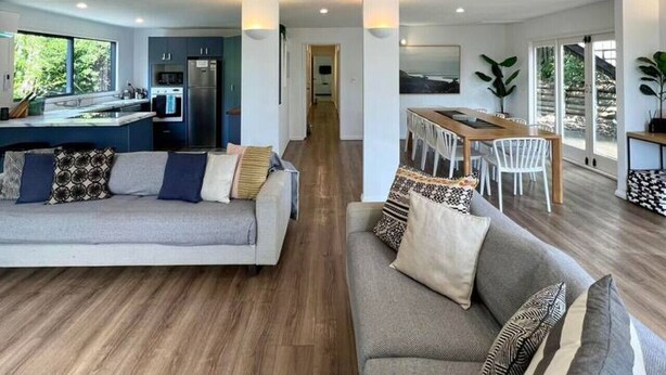 A spacious living area with a sofa and a dining table, a kitchen in the background, and large windows providing natural light.
