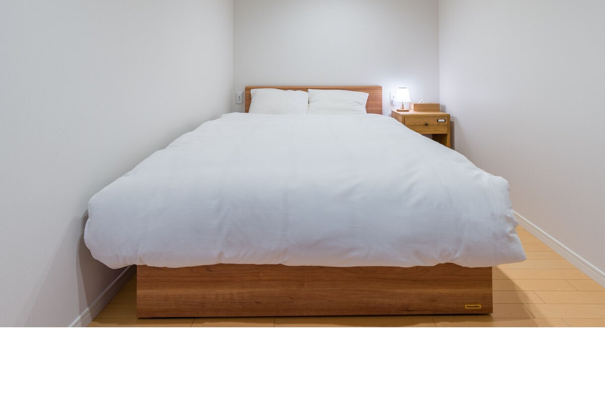 Cama bem arrumada com roupa de cama branca, cabeceira de madeira, mesa de cabeceira com abajur e criado-mudo.