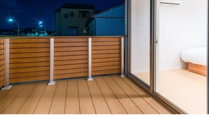 Un balcon avec des balustrades en bois, une porte-fenêtre coulissante donnant sur une chambre à coucher avec un lit, et une vue sur un immeuble en arrière-plan.