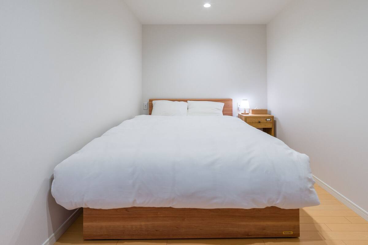 Ein ordentlich bezogenes Bett mit weißer Bettwäsche, ein Holz-Kopfteil, ein Nachttisch mit Lampe und ein Beistelltisch.