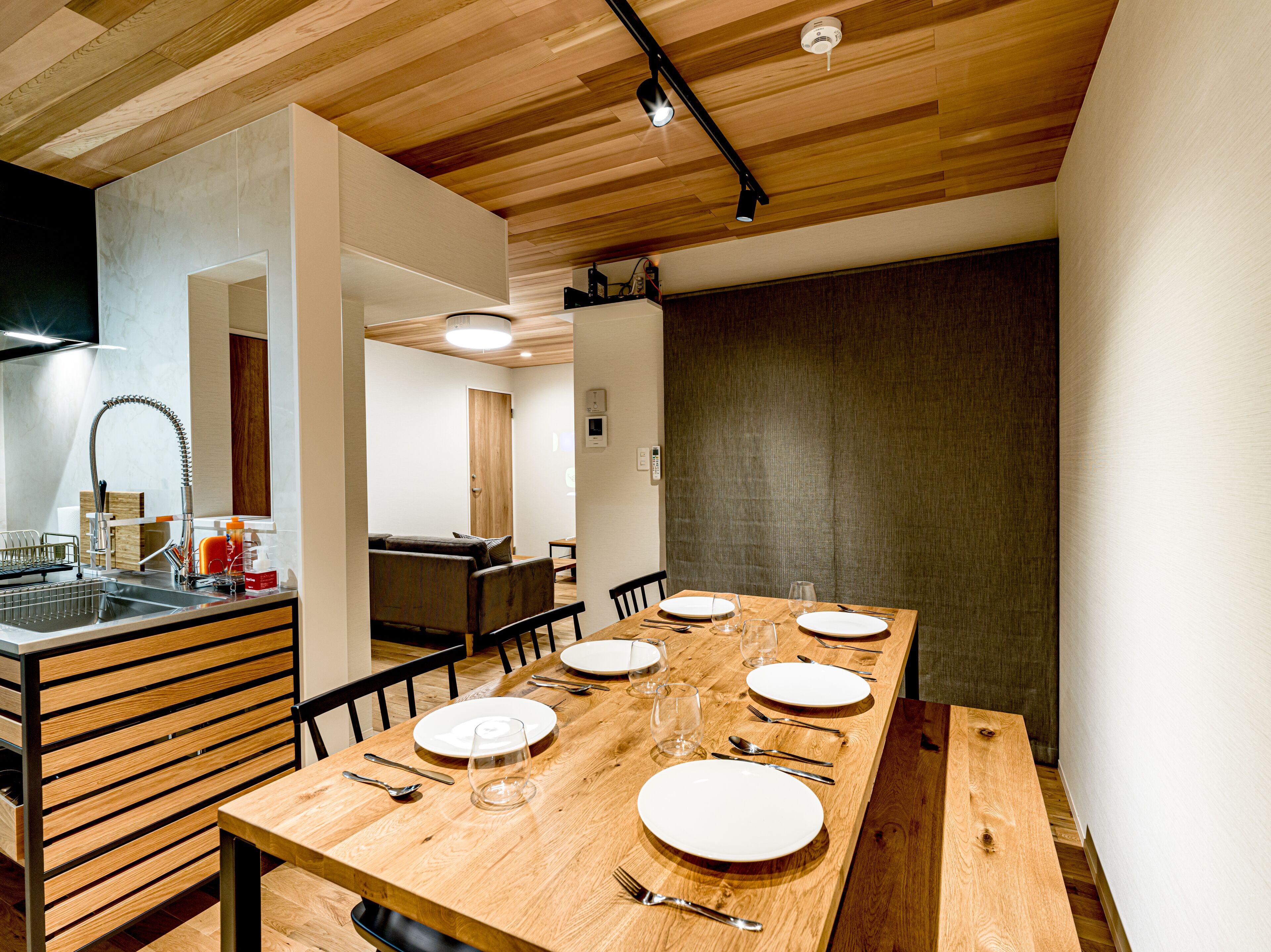 A modern dining area with a wooden table set for six, a kitchen with a sink and stove, and a wooden ceiling.