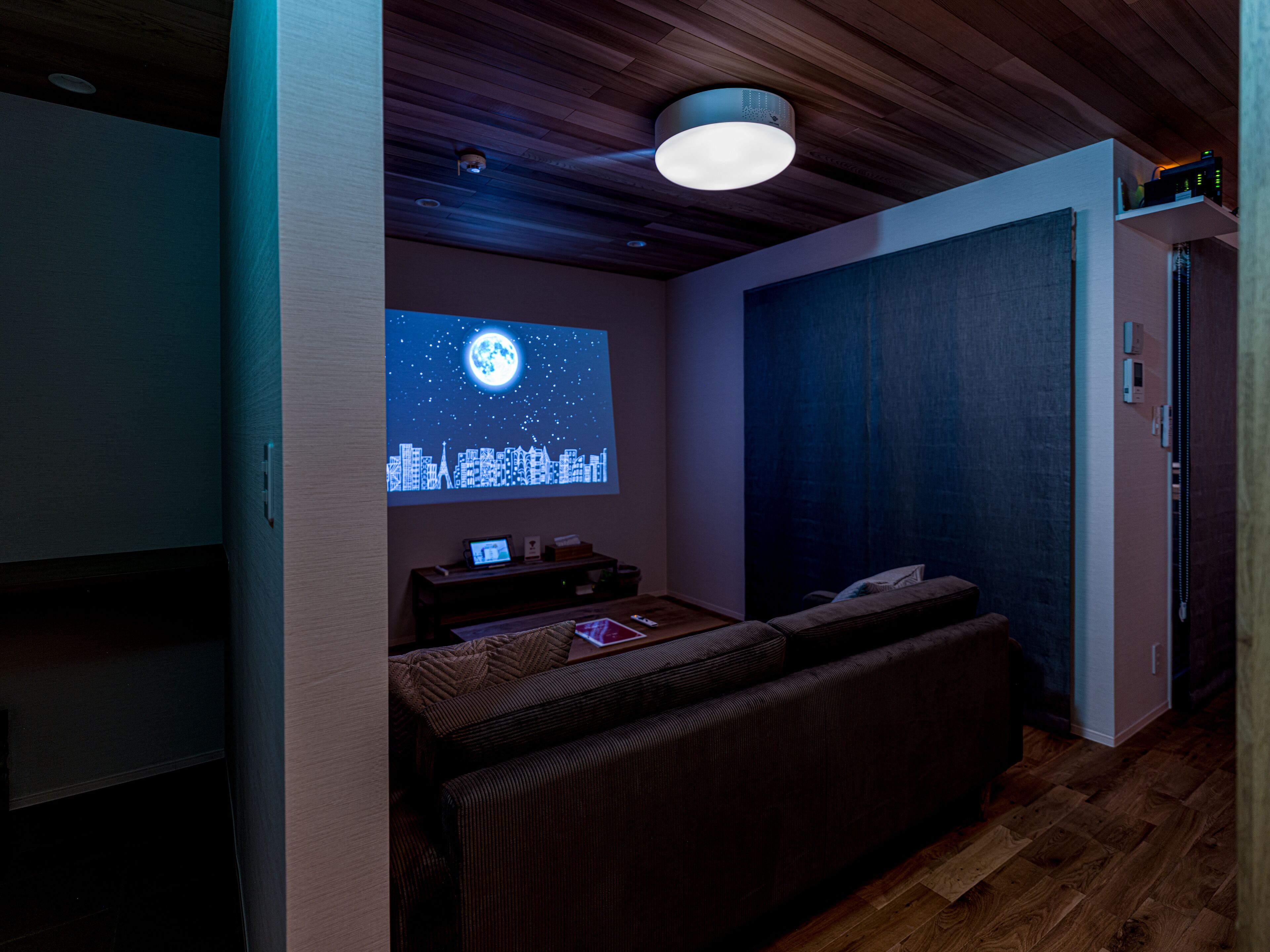 A cozy room with a wooden ceiling, a wall-mounted projector displaying a cityscape, a small table with a laptop, and a sofa.