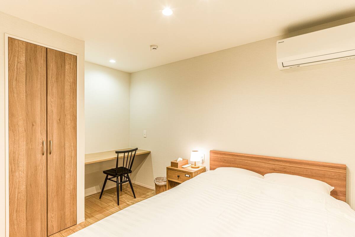 A bedroom with a bed, a desk, a chair, a nightstand with a lamp, and a wardrobe.