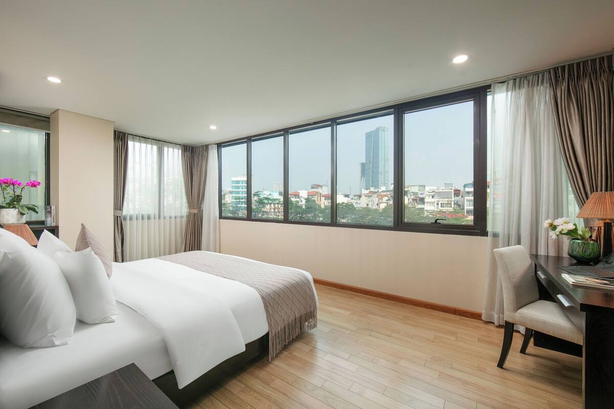 Quarto de hotel com cama grande, escrivaninha com cadeira e vista para a paisagem urbana através de amplas janelas.