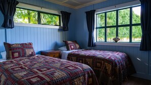 A room with two beds, each with a quilted bedspread, a nightstand, and a window with curtains. - The Jolly Poacher Retreat (Spring Creek)
