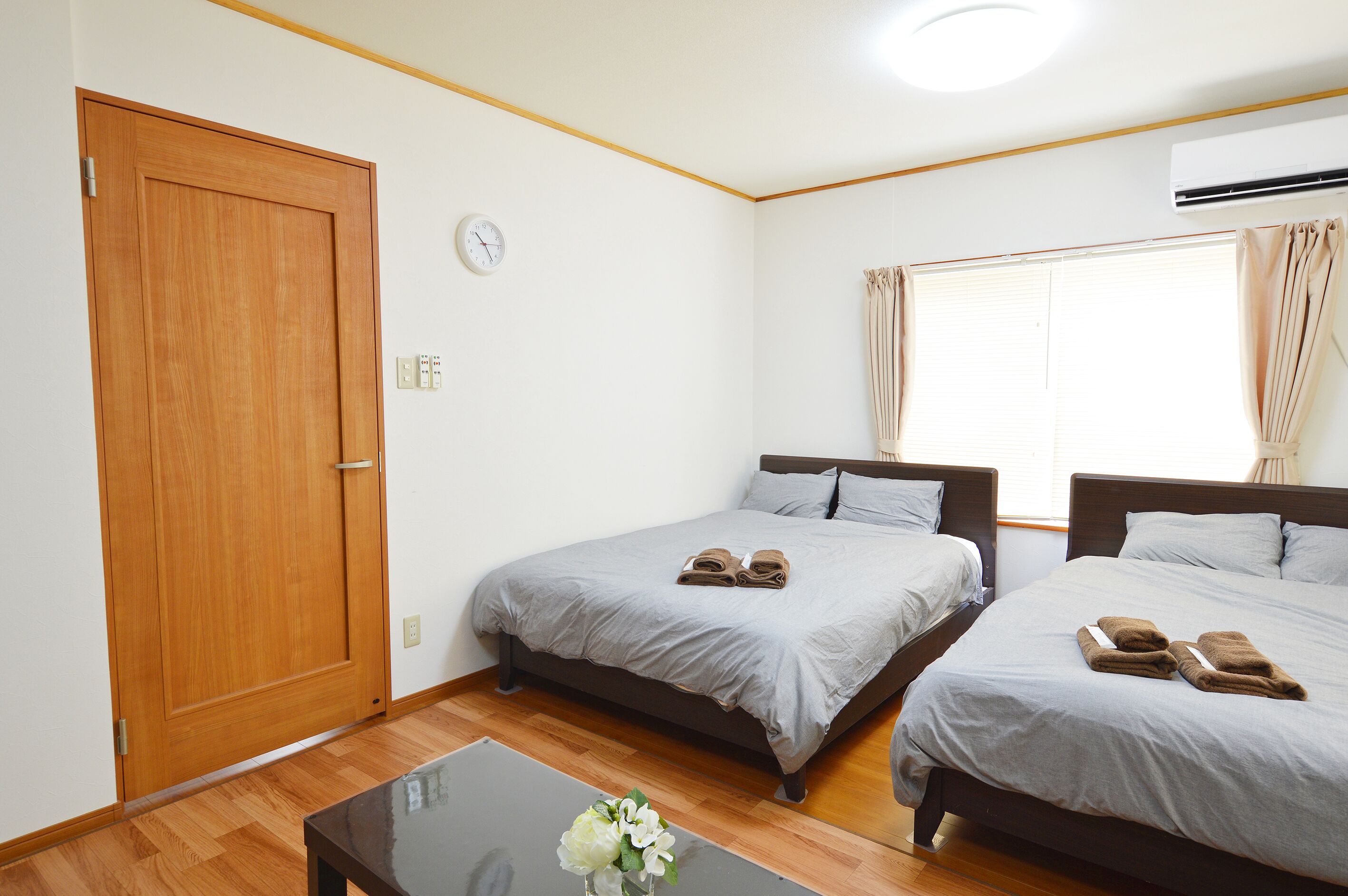 A hotel room with two beds, a wooden door, a clock, an air conditioner, and a window with curtains.