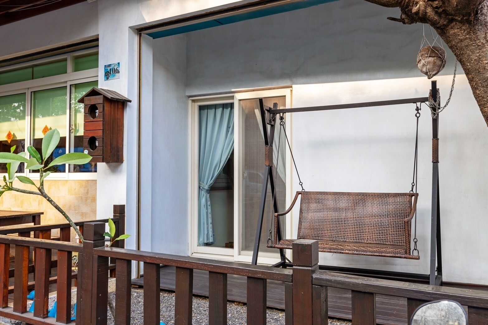 A wooden swing chair hanging from a metal frame, a wooden fence, and a building with a blue awning and a window with a curtain.