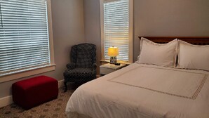 Un dormitorio con una cama grande, dos ventanas con persianas, una silla, una mesita de noche con una lámpara y un reposapiés rojo.