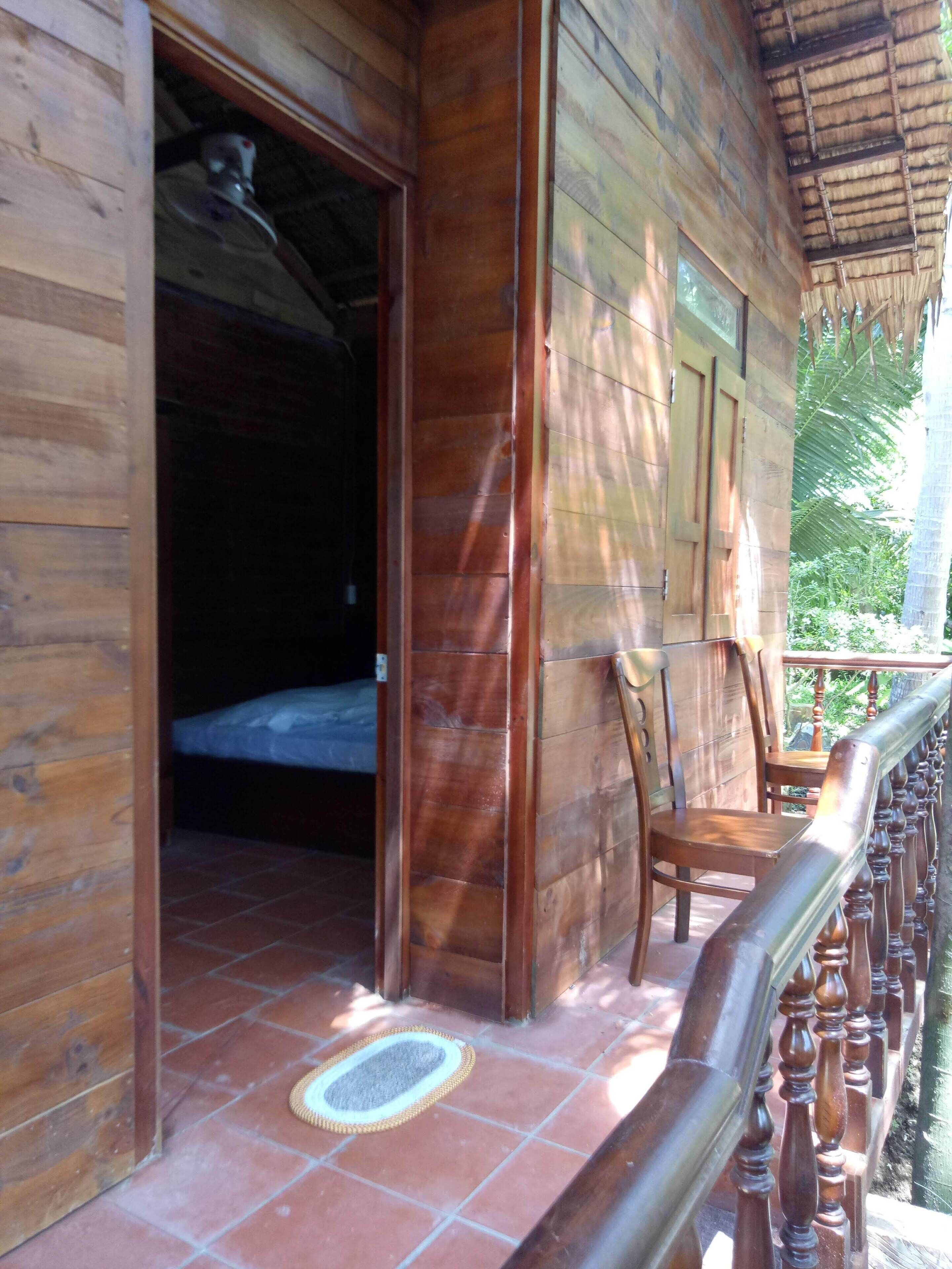 Una cabaña de madera con balcón, una cama a la vista en su interior y suelo de baldosas.