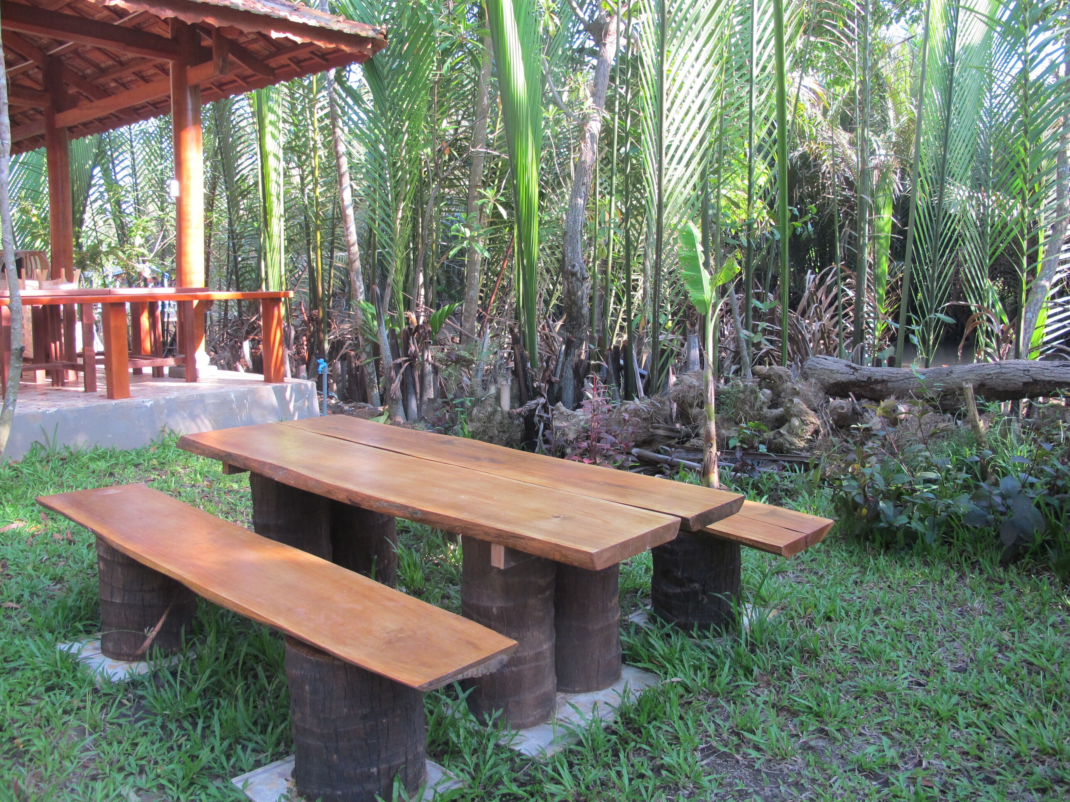 A wooden picnic table with benches, surrounded by lush greenery and a wooden structure in the background.